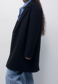 Mujer con un abrigo oversized negro sobre una chaqueta de denim y jeans azul claros, de pie frente a un fondo claro y liso.