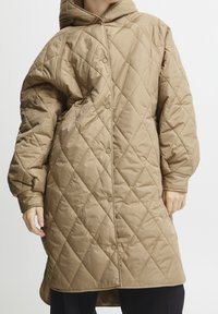 Beige vadderad lång kappa med huva, försedda med diamant-quiltning och tryckknappar. Designad för värme med stora ärmar och en rundad nederkant.