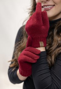 Rote gestrickte Handschuhe mit geripptem Muster, enganliegender Form und einem Stoffetikett am Handgelenk. Getragen von einer Person vor einem hellen Hintergrund.