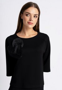 Czarna bluza z trzema ćwierć rękawami; posiada satynową aplikację w kształcie serca z koralikowym wykończeniem na lewym ramieniu. Gładka faktura tkaniny.