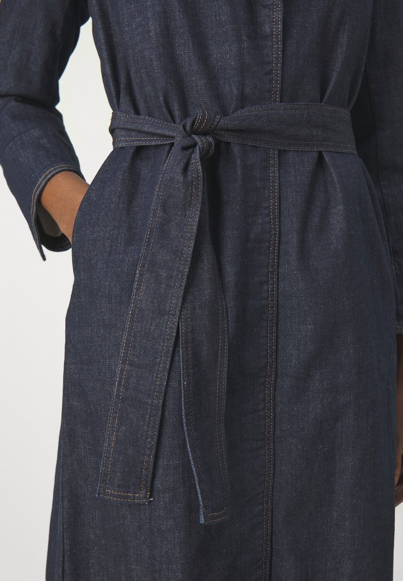 Abito in denim scuro con maniche lunghe, cintura con nodo in vita e dettagli di cucitura sottili. Presenta una chiusura frontale con bottoni e tasche laterali.