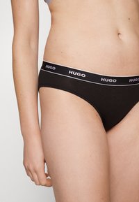 Svarte damestrømper med en myk, glatt tekstur; har en bred elastisk linning med "HUGO"-logo i hvitt.