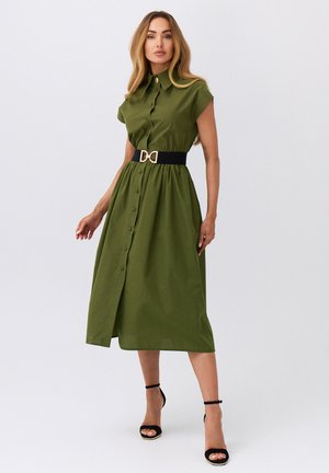 Kobieta stojąca w oliwkowej sukience midi zapinanej na guziki, z krótkimi rękawami typu kapturek, czarnym paskiem ze złotą klamrą i czarnych butach na obcasie z paskiem wokół kostki.