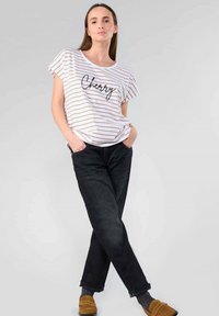Camisa de cortos de manga blanca y dorada a rayas con texto "Cherry", combinada con jeans oscuros y zapatos de tela beige, sobre un fondo gris.