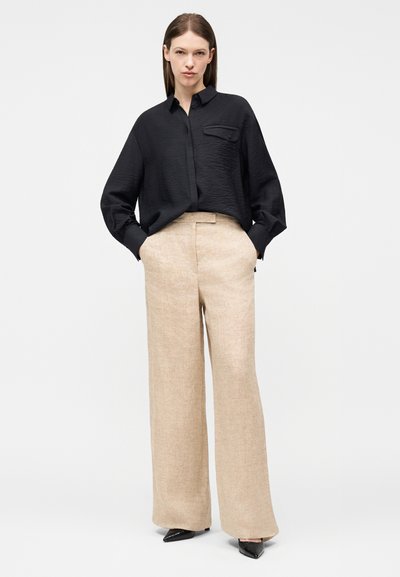 Γυναίκα στέκεται με τα χέρια στις τσέπες, φορώντας χαλαρό μαύρο πουκάμισο με μακριά μανίκια και μπεζ παντελόνια φαρδιάς γραμμής με ψηλή μέση, συνδυασμένα με μαύρα τακούνια.