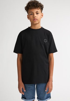 Musta puuvillainen t-paita, jossa on lyhyet hihat, pyöreä kaula-aukko ja pieni logo vasemmassa rinnassa; yhdistetty farkkushortseihin.