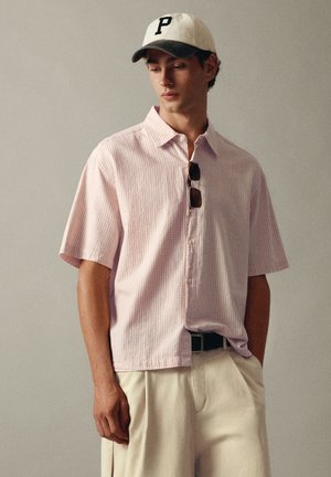 Jeune homme portant une chemise rose clair à manches courtes boutonnée, un pantalon blanc, une ceinture noire, des lunettes de soleil accrochées à la chemise et une casquette blanche avec la lettre P.