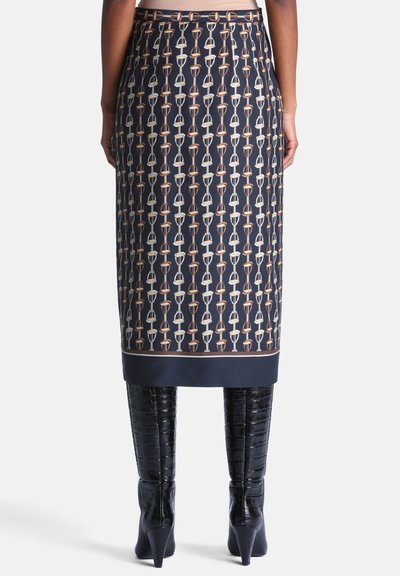 Vue de dos d'une personne portant une jupe bleu marine longueur genou avec un motif répétitif d'étriers et des bottes noires montantes à texture crocodile.