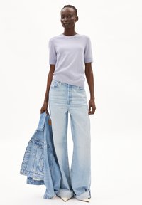 Lichtpaarse korte mouwen trui gecombineerd met wijde lichtblauwe jeans, terwijl een spijkerjack wordt vastgehouden. De outfit heeft een ontspannen pasvorm en zachte texturen.