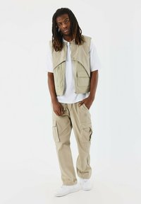 Pantaloni cargo beige e un gilet senza maniche abbinato con tasche frontali. Indossati sopra una maglietta bianca, abbinati a sneakers bianche.