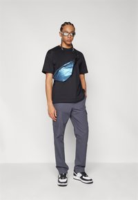 T-shirt noir, pantalons cargo gris et baskets noir et blanc. Une banane bleue brillante portée en travers du corps. Des lunettes fines complètent le look.