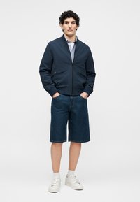 Jeune homme debout, les mains dans les poches, portant une veste bomber bleu marine, une chemise bleu clair, un short en jean foncé, des chaussettes blanches et des baskets blanches.