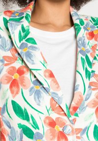 Kwiatowy blezer wykonany z białej tkaniny ozdobionej żywymi kwiatami w kolorach czerwonym, niebieskim i zielonym. Charakteryzuje się kołnierzem i guzikiem.