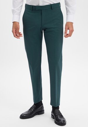 Mann trägt dunkelgrüne, maßgeschneiderte Hose kombiniert mit schwarzen Lederloafern und weißem Hemd, steht vor einem schlichten Hintergrund.