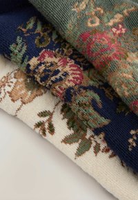 Tre tekstilstykker stablet: en grøn med blomster mønstre, en marineblå med multicolore blomster designs, og en creme med indviklede detaljer.