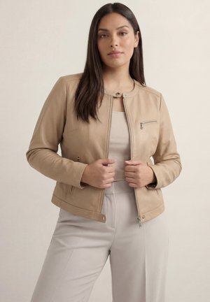 Kobieta o długich ciemnych włosach, ubrana w beżową skórzaną kurtkę na jasnobrązowym topie oraz jasnoszarych spodniach z wysokim stanem, stojąca na tle jednolitego tła.