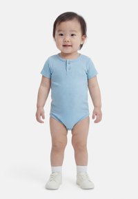 Väikelaps seisab näoga otse kaamerasse, kandes helesinist lühikese varrukaga bodi, valgeid sokke ja valgeid jalanõusid, taust on üleni valge.