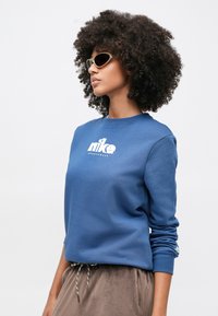 Μπλε φούτερ με λευκό λογότυπο Nike, στρογγυλή λαιμόκοψη και μακριά μανίκια· μοντέλο με γκρι γυαλιά ηλίου και απαλό καφέ παντελόνι.