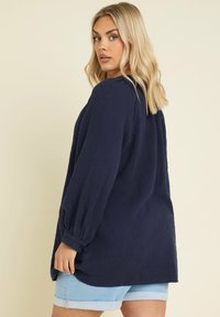 Blusa lunga blu navy, goffrata, con maniche lunghe e polsini abbottonati, vestibilità comoda e spacchi laterali, abbinata a pantaloni shorts in denim blu chiaro.