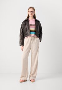 Jachetă scurtă din piele maro, pulover cu dungi roz, pantaloni lărgi bej, pantofi roz slip-on, ochelari cu ramă, fundal neutru.