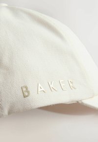 Gros plan d'une casquette en tissu blanc avec une surface texturée et le mot « BAKER » brodé en fil blanc sur le côté.