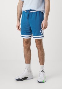 Siniset urheilushortsit valkoisilla yksityiskohdilla ja sivusomisteilla, joissa on verkkokudosta. Yhdistettynä valkoisiin lenkkikenkiin ja nilkkasukkia.