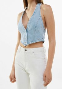 Svetle modrý denimový crop top s hlubokým V-výstřihem, na jedné straně zdobený stříbrnými cvočky a zapínáním na knoflíky v přední části. V kombinaci s bílými kalhotami s vysokým pasem.