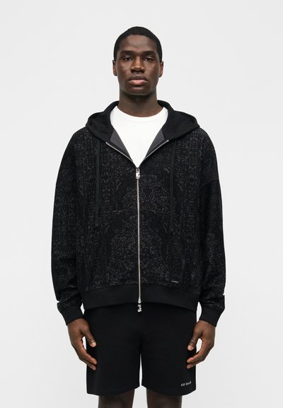 Bărbat purtând o hanorac negru cu fermoar și model peste un tricou alb, împreună cu pantaloni scurți negri, stând în fața unui fundal simplu și deschis la culoare.