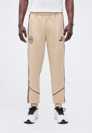 Pantaloni sportivi beige chiaro con loghi neri BVB 09 e Puma King, indossati con sneakers bianche e nere su una persona in piedi.