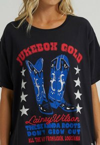 Camiseta negra de gran tamaño con un gráfico de botas de vaquero azules con acentos blancos, texto rojo y estrellas blancas dispuestas en un arreglo circular.