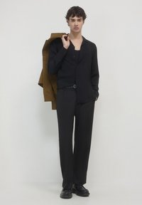 Schwarzes Hemd mit Knöpfen über einem schwarzen Top, kombiniert mit schwarzen Hosen. Eine braune Jacke ist über eine Schulter geworfen. Schwarze Schuhe runden den Look ab.