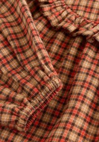 Tissu écossais marron avec des carreaux rouges, noirs et crème ; texture douce ; présente un élastique froncé sur les bords et des détails froncés.