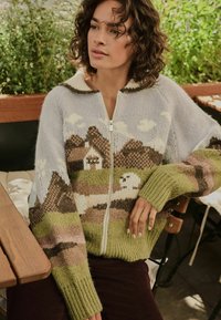 Knit zip-up sweater featuring mountain and house patterns in pastel colors. Made of soft yarn with a ribbed green cuff and collar accents.