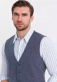 Texturovaná námořnická vesta přes světle modrou a bílou pruhovanou košili, se  špicatým výstřihem, pěti knoflíky a přiléhavým střihem.