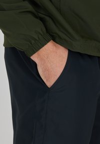 Mörkgrön lättviktsjacka med elastiska ärmar, tillsammans med marinblå byxor som har en sned sidoficka. Slät, matt textur.