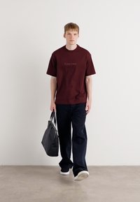 Maroon t-shirt with Calvin Klein logo, paired with dark jeans and black bag. Model is standing on a neutral background, wearing white sneakers.