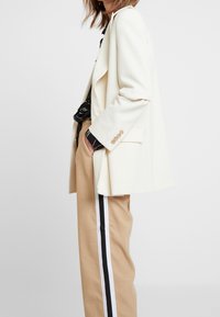 Blazer crème à coupe décontractée, avec des boutons de couleur claire et des poches latérales. Associé à un pantalon beige avec des bandes latérales noires et blanches.