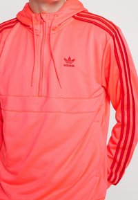 Hoodie Adidas rose vif avec un col zippé, présentant des accents à trois bandes rouges le long des manches et un logo sur la poitrine. Texture lisse.