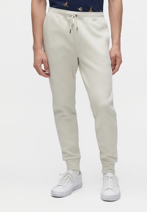 Homme portant un pantalon de jogging écru avec taille à cordon et des baskets blanches, debout devant un fond uni.
