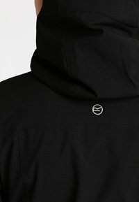 Veste à capuche noire vue de dos montrant un petit logo rond argenté centré sous la couture de la capuche.