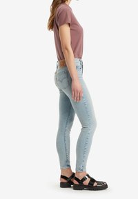 Mujer con jeans ajustados azul claro, camiseta de manga corta color malva y sandalias negras con suela gruesa, de pie de lado sobre un fondo blanco.