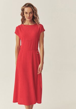 Kobieta o falowanych blond włosach ubrana w teksturowaną czerwoną sukienkę midi z krótkimi rękawami i bocznym detalem z klamrą, stojąca na tle jednolitego jasnego tła.