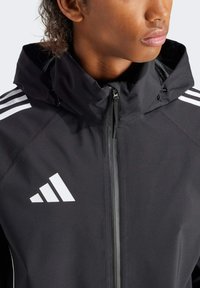 Schwarze leichte Jacke mit Reißverschluss, hohem Kragen und weißem Dreistreifen-Design auf den Schultern. Verfügt über eine glatte Textur und ein Logo-Detail.