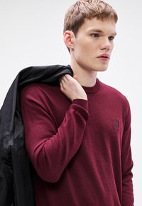 Μπορντό πλεκτό πουλόβερ με στρογγυλή λαιμόκοψη, ριμπ μανσέτες και ένα μικρό ταμπελάκι με το λογότυπο στο στήθος. Μαύρο μπουφάν περασμένο πάνω στον ώμο.