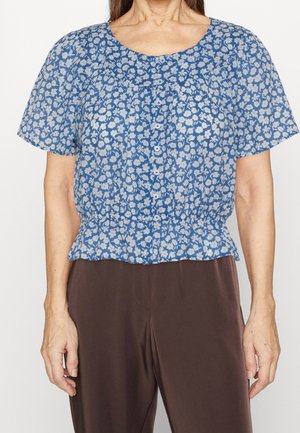 Femme portant un chemisier bleu à manches courtes boutonné avec un imprimé floral blanc et un pantalon marron foncé, vue du cou aux hanches.