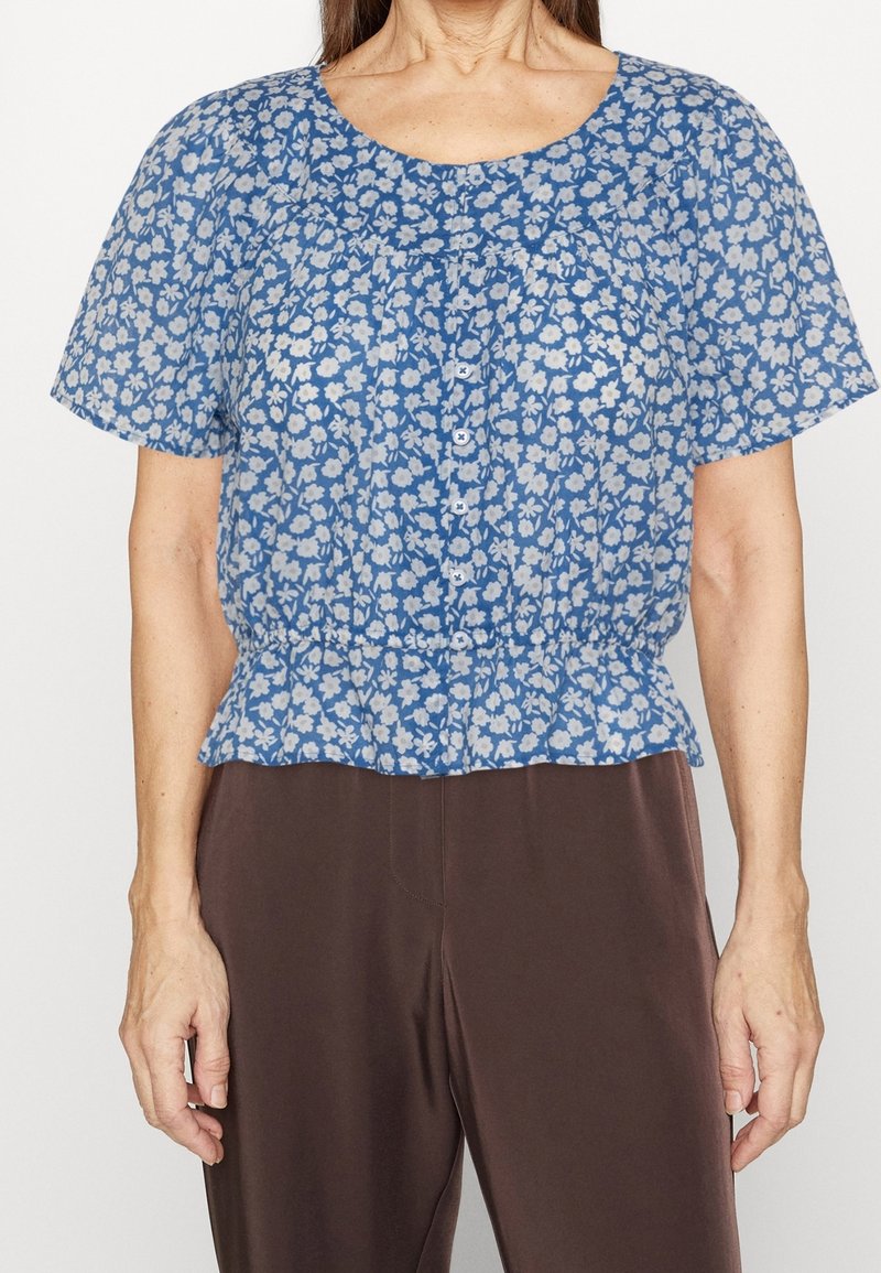 Femme portant un chemisier bleu à manches courtes boutonné avec un imprimé floral blanc et un pantalon marron foncé, vue du cou aux hanches.