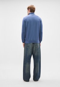 Pulôver azul de malha com gola alta, combinado com jeans de denim azul desbotado e de corte largo. Texturas sólidas, silhueta descontraída.