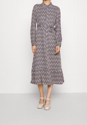 Vrouw draagt een midi-jurk met lange mouwen, knopen en een geometrisch patroon, met een ceintuur in de taille, gecombineerd met crèmekleurige Mary Jane schoenen.