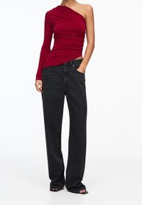 Haut ajusté rouge à une épaule avec un design structuré et une manche longue, associé à un jean en denim noir décontracté.