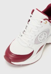 Bílé sportovní tenisky s bordovými akcenty, síťovinovými panely a zlatými očky. Mají kulatou špičku, gumovou podrážku a složité švy.